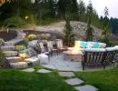 Terraced hillside backyard patio with circular gas fire pit, curved stone seating wall, flagstone path, accent lighting, and stacked stone retaining walls at dusk