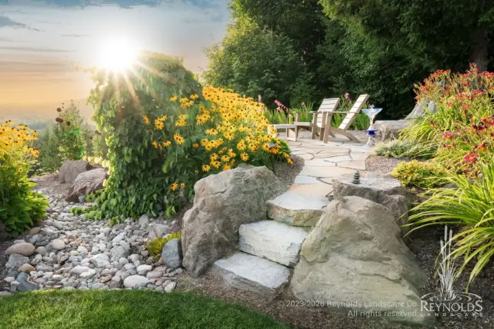 Hilltop view patio with natural stone steps, white Adirondack chairs, dry river rock border, and lush garden plantings bathed in golden sunset light