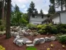 Backyard hillside rockery with scattered natural boulders, bark mulch ground cover, and young ornamental plantings alongside single-story home with mature evergreen backdrop