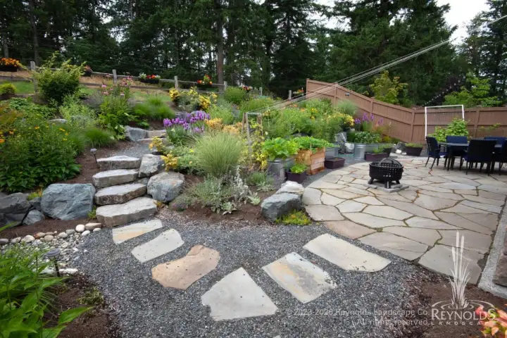 Large flat stone stepping path with gravel surround ascending terraced hillside garden with ornamental grasses, shrubs, and wood fence backdrop