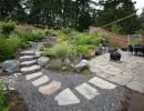 Large flat stone stepping path with gravel surround ascending terraced hillside garden with ornamental grasses, shrubs, and wood fence backdrop