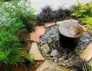 Copper urn bubbling fountain set in a river rock basin with flagstone edging surrounded by ornamental grasses and dark-foliage garden plantings