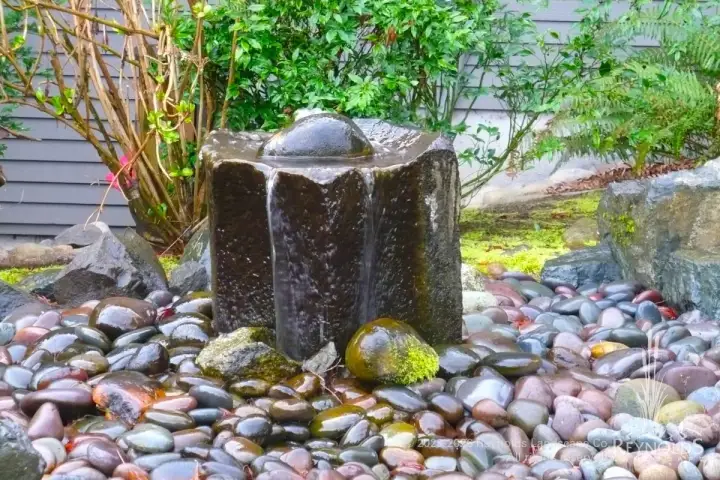 Basalt column bubbling fountain surrounded by river rock and ornamental grasses with Japanese maple accent