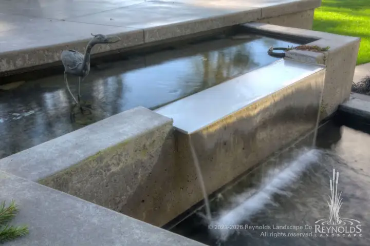 Concrete sheer descent sheet waterfall spilling from a raised rectangular pond into a lower basin on a modern patio