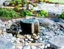Bubbling urn water feature surrounded by river rock and decorative stone with garden border and paver patio backdrop