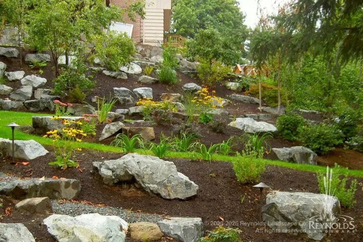 Naturalistic hillside rockery with scattered boulders, winding grass access pathway, bark mulch ground cover, and low-growing ornamental plantings alongside home foundation