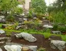 Naturalistic hillside rockery with scattered boulders, winding grass access pathway, bark mulch ground cover, and low-growing ornamental plantings alongside home foundation