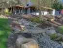 Dry river rock drainage swale with large boulders winding through sloped hillside landscape alongside single-story home