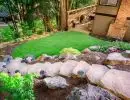 Leveled lawn area carved into wooded hillside with natural boulder slab stairway, solar lighting, lush garden plantings, and deck with pergola above