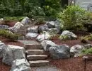 Rustic stone slab steps ascending boulder-lined hillside with wood chip ground cover and mature evergreen tree backdrop