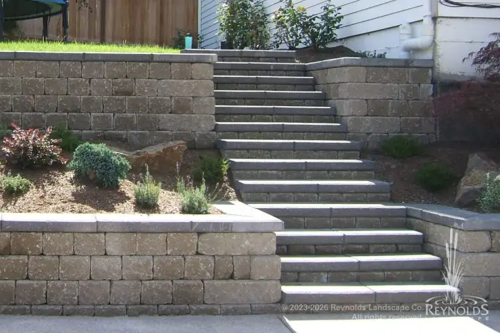 Concrete staircase with block retaining walls on terraced hillside with gravel ground cover and residential home backdrop