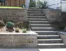 Concrete staircase with block retaining walls on terraced hillside with gravel ground cover and residential home backdrop