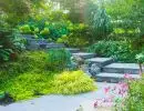 Lush hillside garden with natural stone retaining walls, layered ferns, flowering perennials, and dense ornamental shrubs