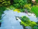 Modern large format paver stairway descending hillside with lush green garden beds, ornamental boulders, and manicured lawn backdrop
